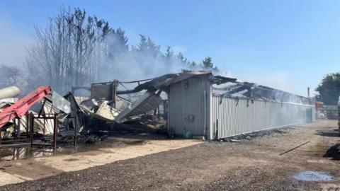 A large storage building which has been involved in a fire. Smoke is billowing out the building. The roof has been burnt and destroyed in the blaze.