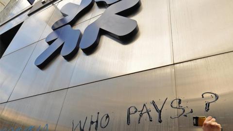 A protestor scribes the words 'who pays' onto the side of an RBS building.