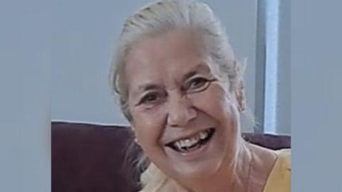 Image of a smiling Helen Holland, a woman with long grey hair tied in a ponytail and wearing a yellow top