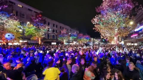 A crowd of people watching the Plymouth city centre light switch-on. There is a blue glow over the crowd and trees are wrapped in colourful Christmas lights.