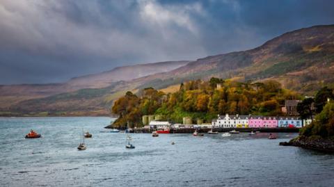 A row of three-storey properties line Portree's harbour. The properties are brightly coloured - white, pink and blue. Behind the buildings is a wooded hill, and bare hills behind that. There are boats anchored in the bay and an orange lifeboat headed towards shore. 