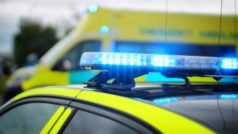 A parked police car with it's blue lights flashing in the foreground. An ambulance is visible behind it. 