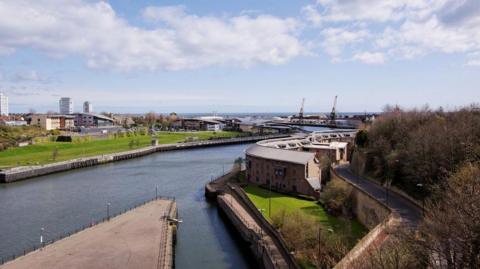 A view of the River Wear with large modern buildings on either side. On the left is a large patch of grass. Further down the river there are two large cranes. The sea can be seen beyond them.