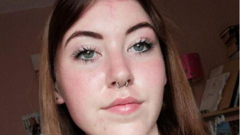 A teenage girl with should length, brown hair. She has a pale complexion and is wearing a nose ring.