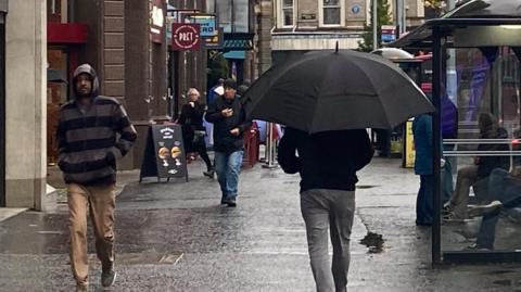 Belfast city centre, man walking away from the camera in grey trousers and black jacket holding a black umbrella. Another man walking towards the camera in tan coloured trousers and a grey and black stripes hoodie  