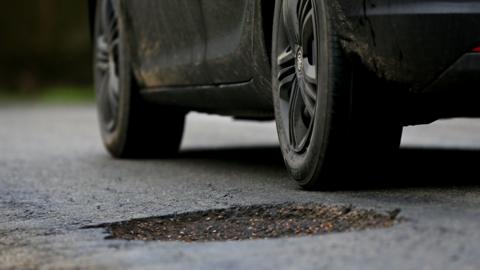 A large pot hole in a tarmac road with a black car driving past.