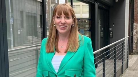 Angela Rayner in Hanley wearing a light green top and trousers over a white top while standing on pavement next to a building with a railing outside