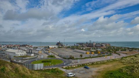 The Europort at Rosslare, it is a clear day with some clouds in the sky. At the port there are vehicles parked. In the horizon you can see the sea. 