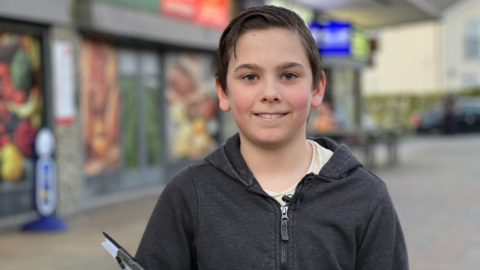 A young boy wearing a zipped-up hoodie stood in front of a Morrisons Local holding a clipboard and paper