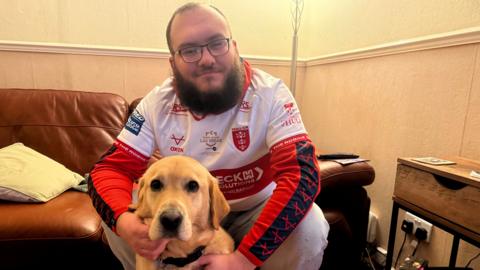Nathan Major is wearing a Hull KR rugby shirt is sitting on a sofa while holding a golden Labrador guide dog gently between his arms. The dog is looking directly toward the camera, resting calmly in front of him in a warm, homely living room.