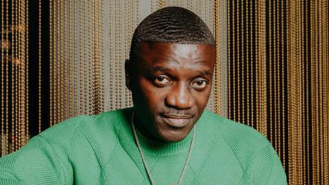 Akon wearing a green top with his hands joined together at the fingertrips. He is wearing a gold necklace as he looks into the camera. The background is shiny gold pearls dropping from top to bottom, as his right elbow is perched on a table.