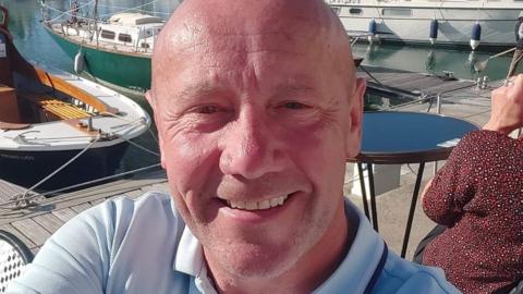 A bald man smiles at the camera. He is sitting next to a marina with small boats behind him