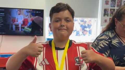 Oliver looking towards the camera with his thumbs up. He is smiling and is wearing a red and white striped shirt. There is a Exeter City FC badge on the right side of the shirt. Behind him is a TV and a woman stood to his right.