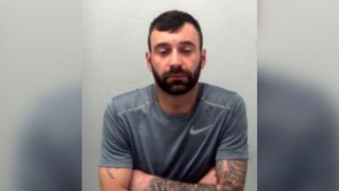 Sidney Kemp has short black hair and a beard. For this picture, taken in police custody, he is wearing a grey T-shirt and looking downwards.