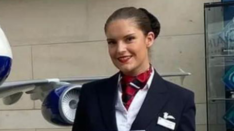 A woman in an air steward's uniform stands next to a statue of a British Airways aeroplane. The woman has dark brown hair and she is wearing a navy blue skirt suit, a white shirt, and a red and white scarf. She is also holding a certificate. The aeroplane statue is mostly white, with red and blue designs on it.