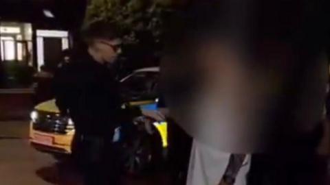 A male police officer with light blonde hair and glasses takes the right hand of a man, who is wearing a white t-shirt, and places it in handcuffs. A police patrol car is visible behind the officer. 