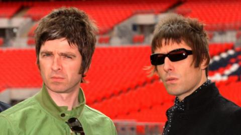 Noel and Liam Gallagher, standing in a stadium with red seats in the background. Noel, on the left, wears a green jacket with sunglasses hanging from the collar. Liam sports a black jacket and is wearing with sunglasses.