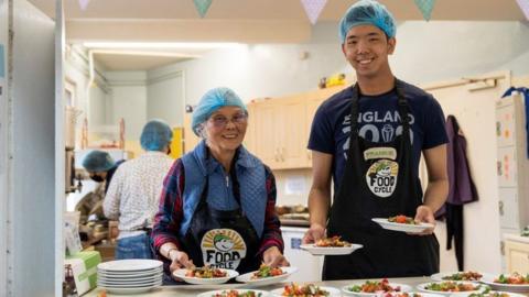 Pair of smiling volunteers with blue hair nets hold plates of food at a food counter with kitchen in background