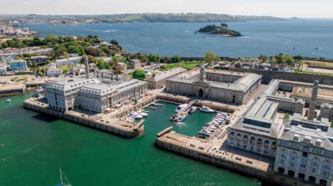 An aerial photo of Royal William Yard on a sunny day. There are a few small boats moored in the harbour. The buildings are granite and there is a formal square in the centre which has a tower on its entrance gate. The sea is in front and behind the promontory, there is an island close in and the coastline continues in the background.