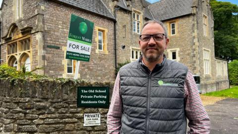 Estate Agent Will Parfitt smiling and wearing checked red and blue shirt with a black puffer jacket. Behind him is a stone wall and behind that is a large brick house