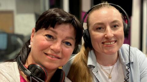 Nataliia Revkova and Nadiia Hoian smile at the camera. They are sat next to each other in a radio studio. Nataliia has dark hair that is tied up and she has a fringe. Headphones rest around her neck. Nadiia has long blonde hair that is tied up and she wears her headphones.