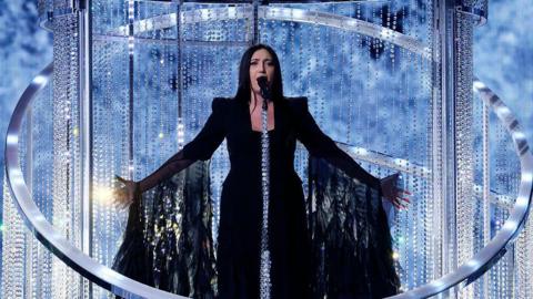 Israel's Eurovision 2025 entrant, Yuval Raphael, sings on stage surrounded by giant hoop chandeliers at the contest in Basel, Switzerland in May. She is wearing a dark dress which has long, frilly sleeves extending to the floor. 