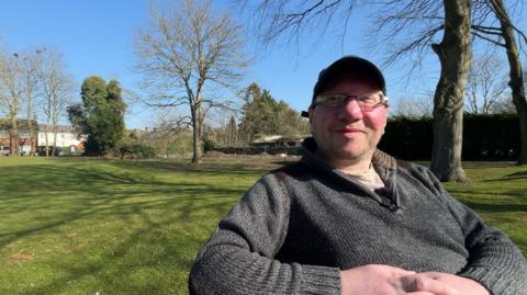 A man sits on a bench in a leafy park. he's wearing a grey jumper and a cap and glasses.