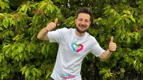 Mitchell Elliott in a white t-shirt that has the logo of Echo on it, in the shape of a heart. He is smiling and giving two thumbs up while stood in front of a large, leafy hedge.