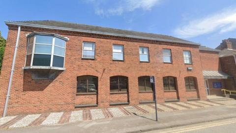 A streetview image of the police station, which is a two-storey red brick building with a number of arched windows. 