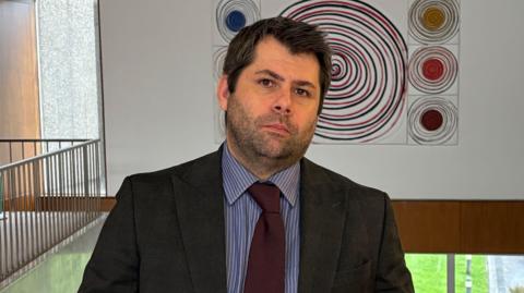 Leigh Frost has short black hair and stubble and wears a dark suit, navy blue lined shirt, and maroon tie. He is standing on top floor of County Hall with a piece of artwork and large windows behind him.