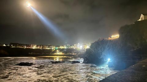 A bright helicopter torch shining on rocks and cliffs. There is water below and lit up buildings in the background.