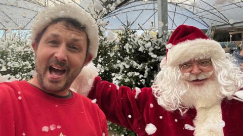 Combe Garden Centre manager James Trevett cheers as he stands next to Father Christmas in front of Christmas trees at the shop. Fake snow made from bubbles is coming down from the sky. Mr Trevett is wearing a red jumper and a Christmas hat.