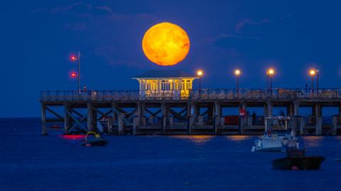 A large bright orange moon in the sky over the pier at Swanage. The moon is perfectly lined up with a small shelter that has lights on that make it look like the moon is inside. There are three boats in the water in front of the pier and there is a row of street lights on the pier. Both the sky and sea are a dark blue. 