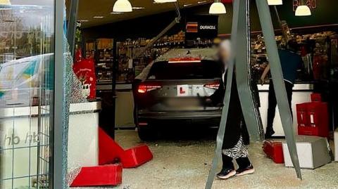 A grey car with an L plate on the back in the middle of carnage, surrounded by smashed glass, tumbled boxes, and hanging metalwork as a woman in a headscarf walks by inside the shop