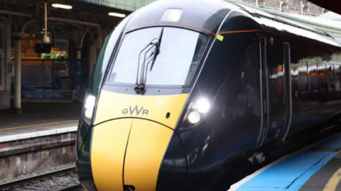 A Great Western Railway (GWR) train arrives at a station. It has yellow front panel and a dark blue body. A blue strip can be seen on the platform. 