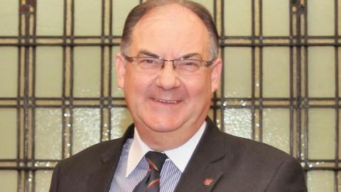 Geoffrey Corkish smiling. He is wearing glasses and a dark suit with a stripy tie.