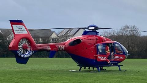 The red and blue Devon air ambulance is stationary on a flat area of grass. A paramedic is climbing in it through an open door.