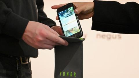 A hand holding a mobile phone just above a grey rectangular wallet, which has a green logo reading "YONDR" in capital letters. The wallet is being held open by a second person who is dressed in black.