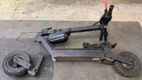 Broken e-scooter pictured on the floor of a repair shop in Aberdeen. The scooter is black with one pink handle. The back wheel and connecting fitting is broken and completely detached from the body of the scooter