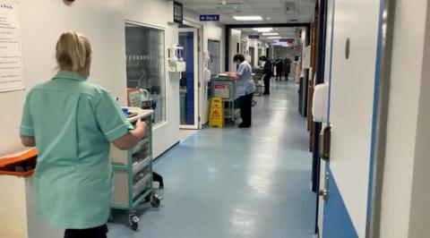 Two women in hospital uniforms at work in a blue-floored medical corridor with white walls and a number of objects lined up against the left hand wall.