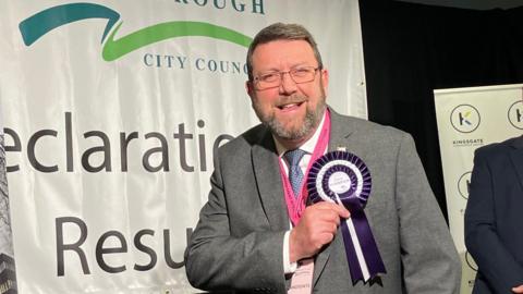 Chris Harper with a beard and glasses wearing a grey suit. He is touching his purple and white rosette with his name on it. A Peterborough City Council sign saying "Declaration Results" is in the background.  