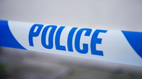A stock image of some police tape with blue writing on a white background. The background is blurred with no identifiable features.