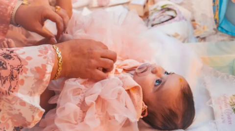 Baby Sophia lies down in a bed. She is wearing pink baby clothes. She sucks a dummy and looks up to the ceiling. A pair of female hands corrects her clothes. The woman wears a gold bracelet and rings.