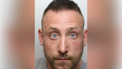 A police mugshot of a man with short dark hair, a short beard and a grey top.