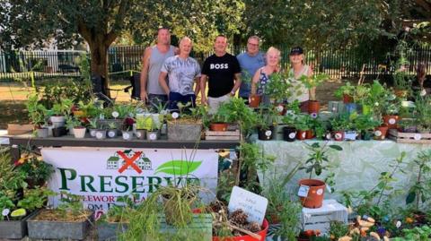 A group of six people (four men, two women) stand behind tables of plants at a plant sale to raise money for the Preserve Littleworth Lane Fields campaign. A banner on one of the tables says 'Preserve Littleworth Lane Fields