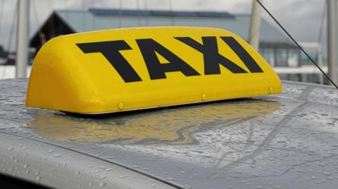 An unlit yellow taxi light on the top of a car on a grey, wet day in the British Isles.