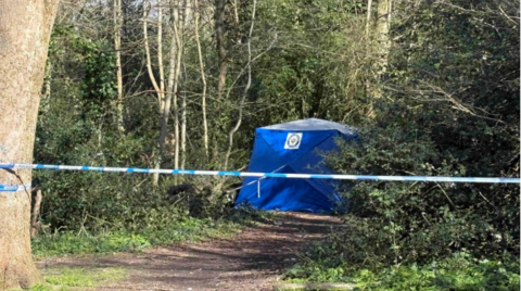 A police cordon attached to a tree in the middle of a wooded area. A blue police tent can be seen further down the footpath next to some trees.