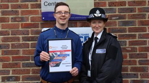 Luke Newton standing next to a female police officer. He has short dark hair, glasses and is wearing a blue shirt. He is holding a certificate with his name on that says "shortlisted". The female officer next to him is wearing a white shirt, black tie, a black police jacket and a police hat. Both of them are looking at the camera and smiling. 