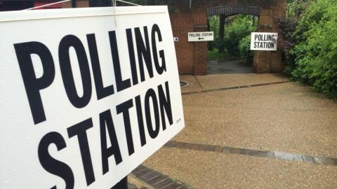 A generic image of a white sign with black lettering for a polling station. In the background there is an arched entrance to a building's garden with further signs pointing to a polling station.