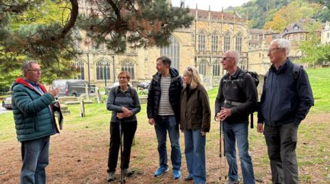A group of two women and three men are listening to a man talking outside. The man talking is wearing a blue puffer jacket and has a hand raised as he describes something. A large church building is behind the group. 